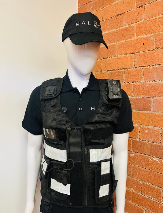 A mannequin wearing a black tactical vest over a black polo shirt, with a black cap featuring the HALOS logo. The vest has reflective white stripes and a mounted body camera on the shoulder, set against a split white and orange brick wall background.